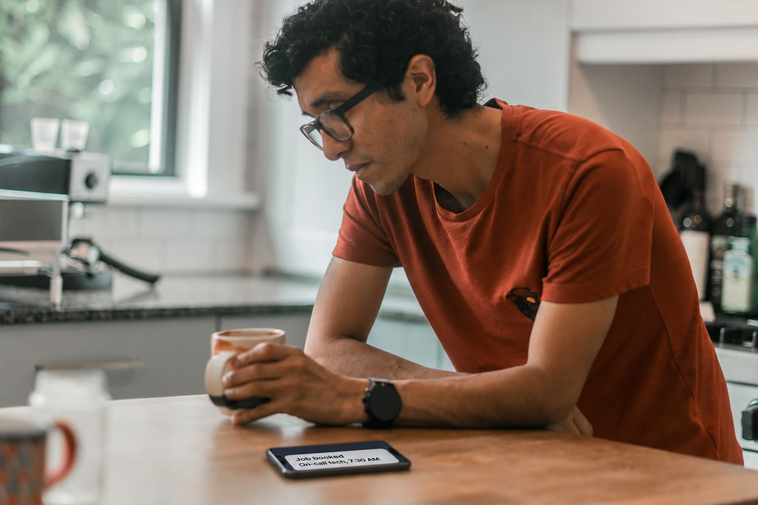 A man with his morning coffee at the kitchen counter, his phone face up showing a job booked overnight