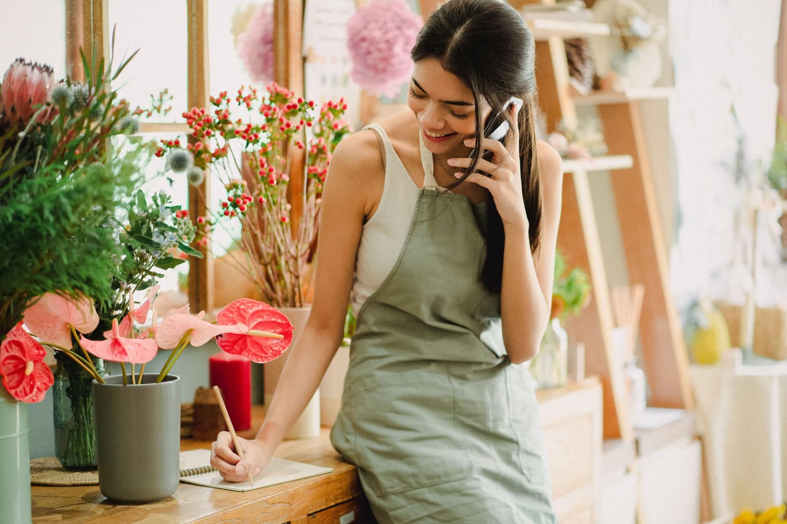 A florist on a phone call at her work table, writing a note as she listens