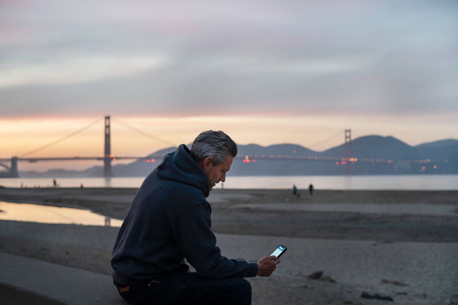 California two party consent: a man checking his phone on a San Francisco beach with the Golden Gate Bridge behind him