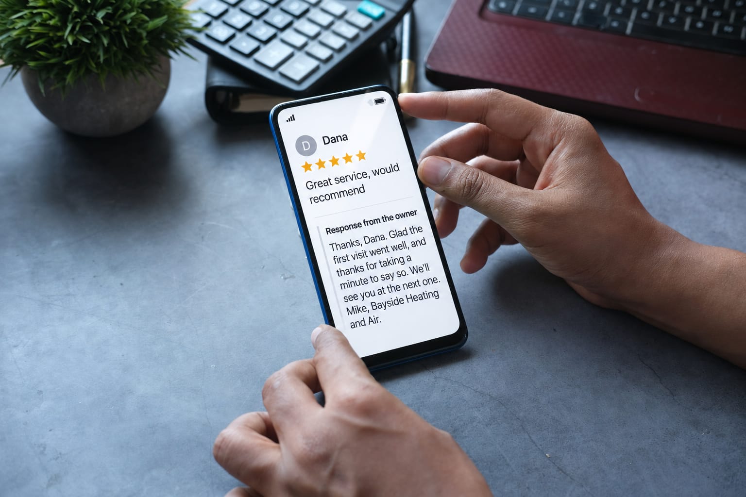 A phone on a desk showing a five-star review and the owner's short reply thanking the customer by name