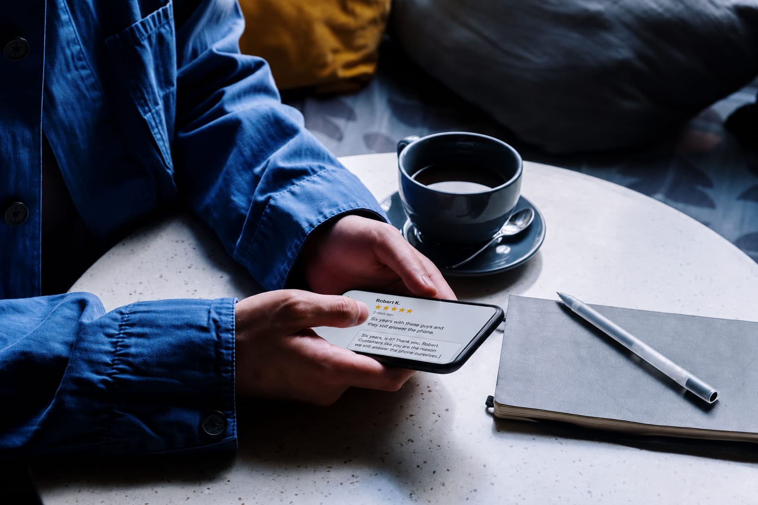 A phone beside a cup of coffee showing a five-star review from three days ago and the owner's reply being typed