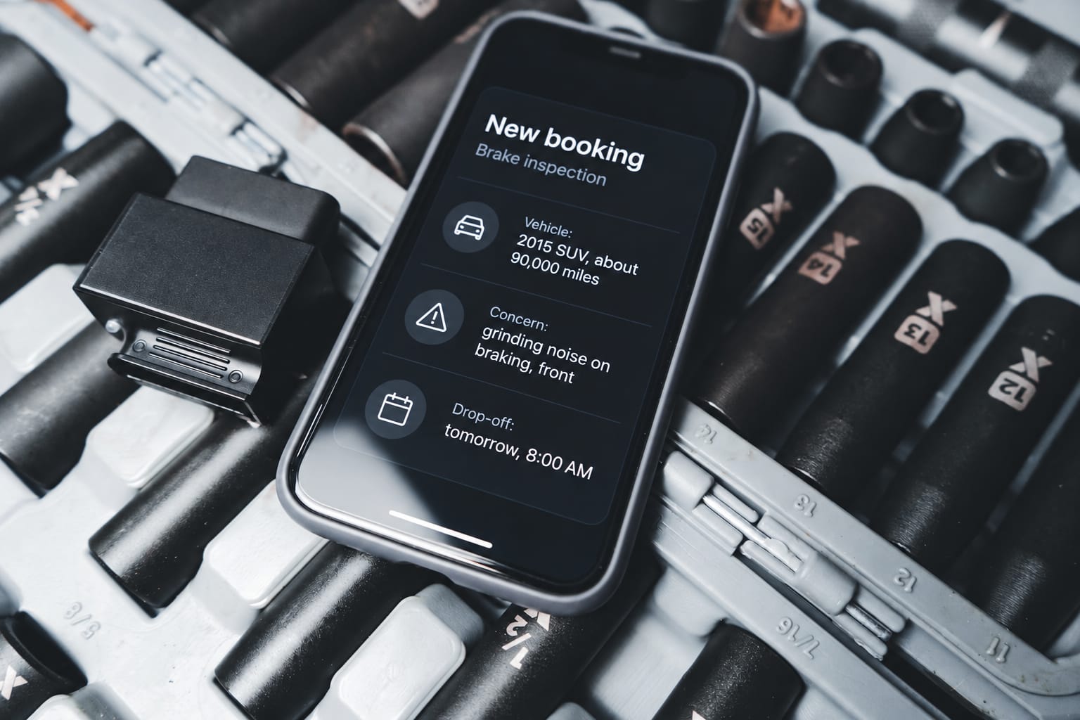 A phone on a socket set in the workshop showing a booked brake inspection: vehicle, concern and drop-off time