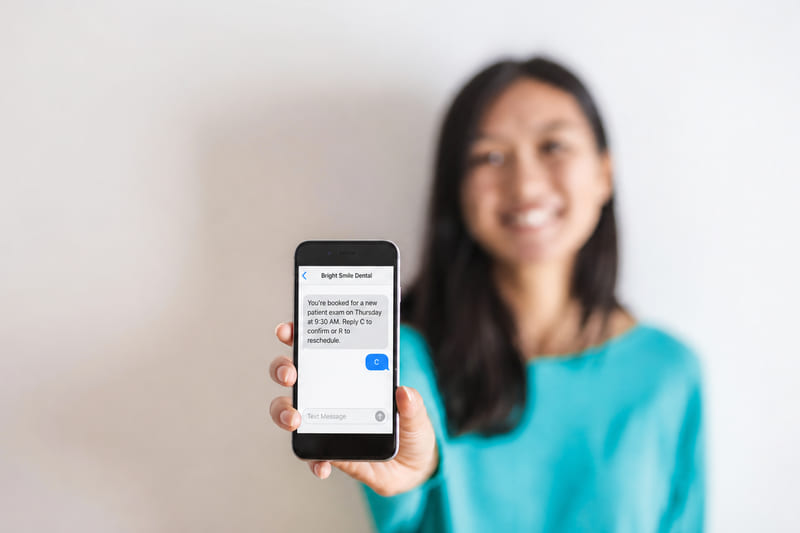 A smiling woman holds up her phone. A text from Bright Smile Dental reads: You're booked for a new patient exam on Thursday at 9:30 AM. Reply C to confirm or R to reschedule. She has replied C.