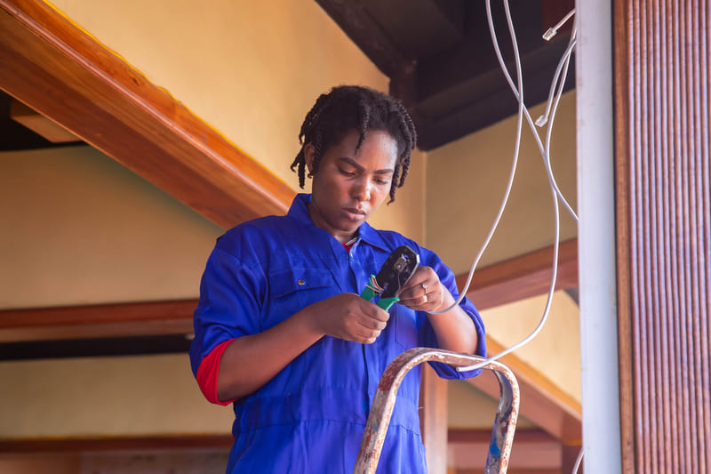 An electrician in blue overalls stripping a wire at the top of a ladder indoors.