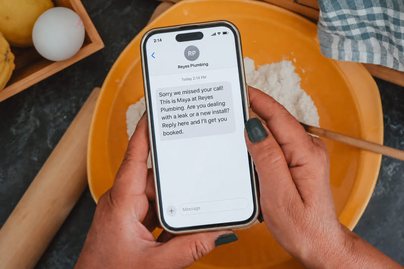 Hands holding a phone in a kitchen, showing a text from Reyes Plumbing that begins: Sorry we missed your call!