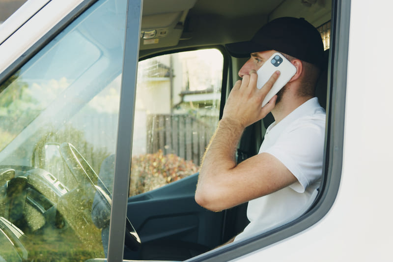 A man in a cap on a phone call in the driver's seat of a white work van.