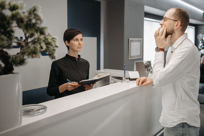A dental receptionist listening to a patient who holds his jaw at the front desk.