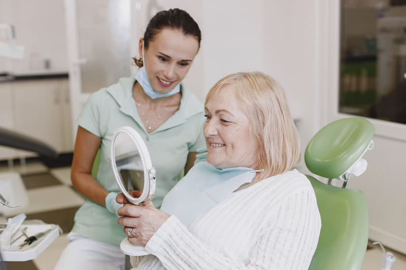A dental hygienist holding a mirror while a smiling patient checks her teeth in the chair.