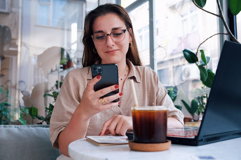 A woman reading her phone at a cafe table, a cold brew and a laptop beside her.